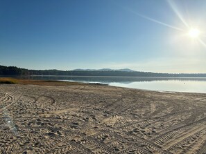 Una playa con surcos de neumáticos que conducen a un lago tranquilo, rodeado de árboles y montañas distantes.