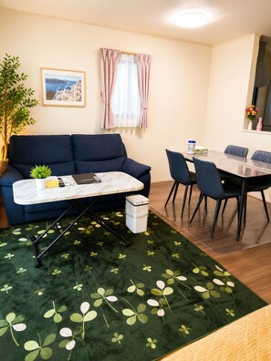 Een knus woongedeelte met een bank, een klein tafeltje, een eettafel met stoelen, een raam met gordijnen, een potplant en een lijst met foto aan de muur.