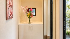 A hallway with a white cabinet, a vase with flowers, a framed picture, and a mirror reflecting the outside. - 2-Min Walk to Koya Station | Brand-New 4BR Private House | Free Parking & Wi-Fi (足立区)
