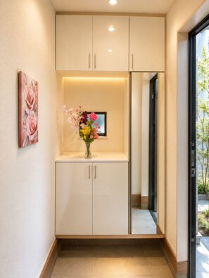 A hallway with a white cabinet, a vase with flowers, a framed picture, and a mirror reflecting the outside. - 2-Min Walk to Koya Station | Brand-New 4BR Private House | Free Parking & Wi-Fi (足立区)
