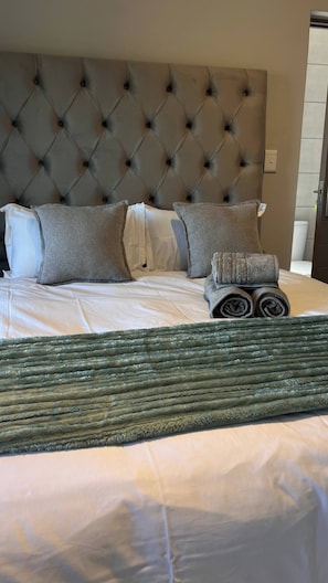 A bed with a tufted headboard, grey pillows, a rolled-up towel, and a green textured throw. - Luxury Apartment Overlooking the River (Johannesburg)