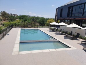 A rectangular pool with a metal ladder, surrounded by a paved deck. There are lounge chairs and umbrellas, and a building with large windows is adjacent to the pool. - Luxury Apartment Overlooking the River (Johannesburg)
