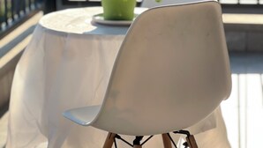 Un balcón con una mesa cubierta por un mantel blanco translúcido, una planta verde en maceta y una silla blanca con patas de madera.