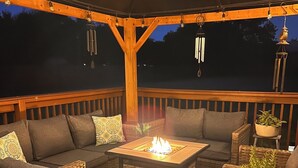 An outdoor deck with wicker furniture, a central fire table, and potted plants.