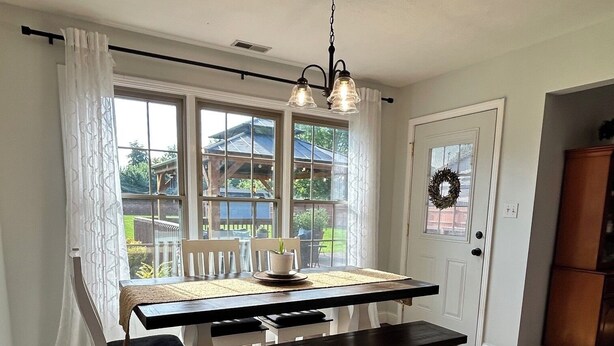 A dining area with a table, chairs, a chandelier, and a view through large windows.