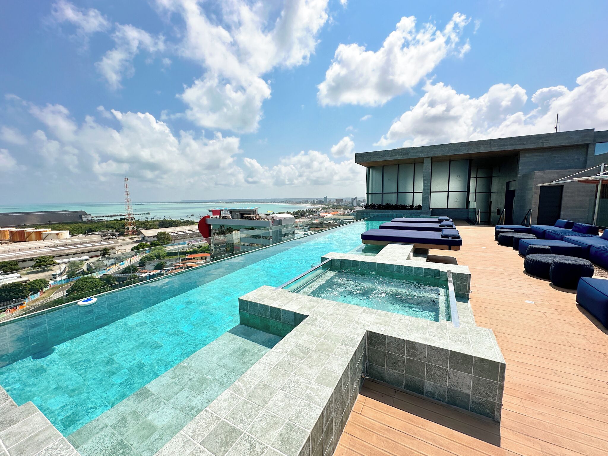 Una piscina infinita en la azotea con vistas al océano, mobiliario de descanso y una estructura de edificio moderna.