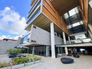 A modern building with 'EIQ' and 'RUCA' signage, outdoor seating, and a reflective glass facade. - Edifício New Time (Maceió)