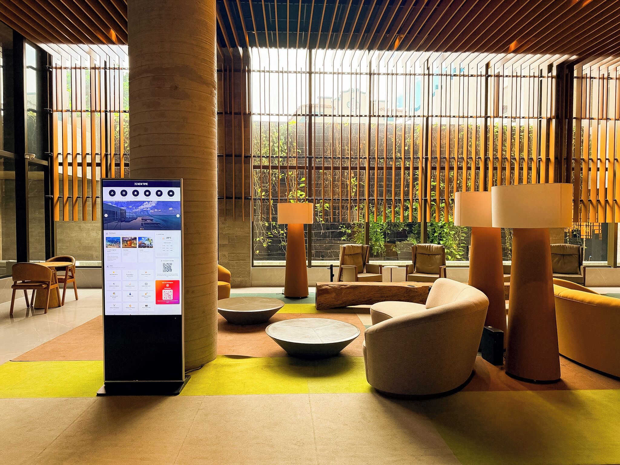 A modern lobby with a digital information kiosk, comfortable seating, and large windows with a view of greenery.