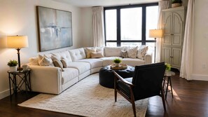 Ein Wohnzimmer mit einer Eckcouch, einem runden Couchtisch, einem schwarzen Sessel, einem weißen Teppich und einem großen abstrakten Gemälde an der Wand.