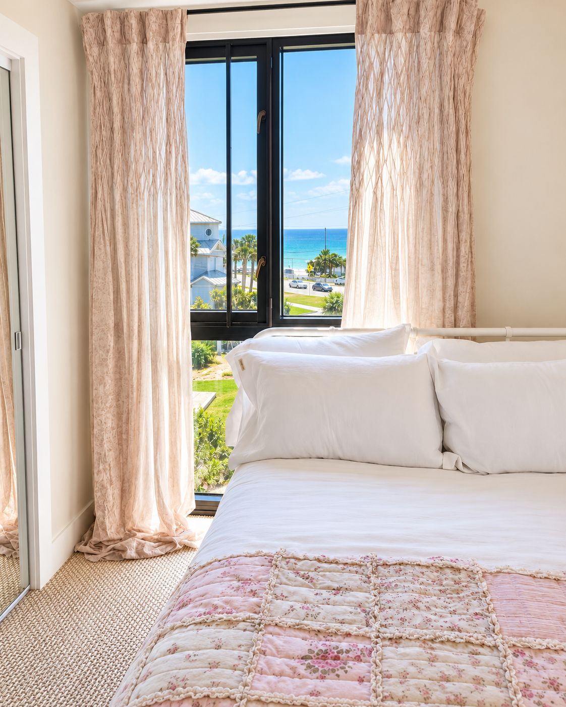 A bedroom with a large bed, white bedding, and a view of the ocean and palm trees.