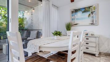Un coin repas avec une table blanche, des chaises en bois, un buffet blanc et un tableau représentant une scène de plage.
