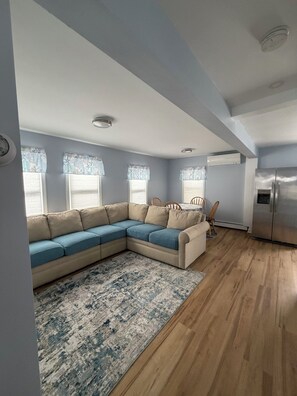 A living room with a sectional sofa, a wooden floor, and a kitchen area with a refrigerator.