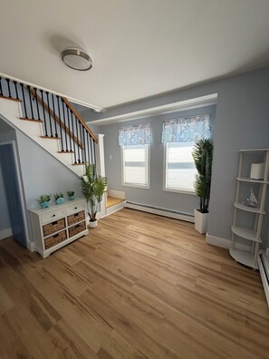 Ein geräumiges Zimmer mit einer Holztreppe, großen Fenstern mit blauen Vorhängen und einem weißen Beistelltisch mit Körben.