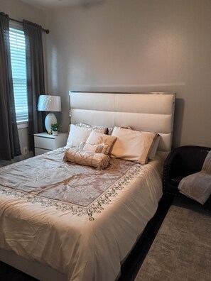Una cama bien hecha con cabecera, una mesita de noche con lámpara y una silla.
