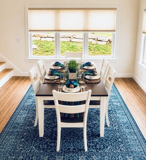 A dining area with a table set for six, white chairs, a blue patterned rug, and a view of a stone wall and grass through large windows.