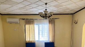 Un salón con piso de baldosas, dos sillones azules, un sofá oscuro, una mesa de centro de madera, una araña y una ventana con cortinas.
