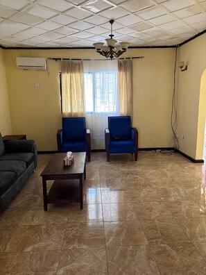 A living room with a tiled floor, two blue armchairs, a dark sofa, a wooden coffee table, a chandelier, and a window with curtains. - BLM Lodge (Monrovia)