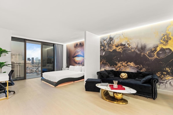 A modern hotel room with a bed, a black sofa, a round coffee table, and a large abstract mural on the wall.