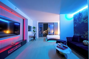A modern hotel room with a large flat-screen TV, a bed with a view of the city, a sofa, and a coffee table.