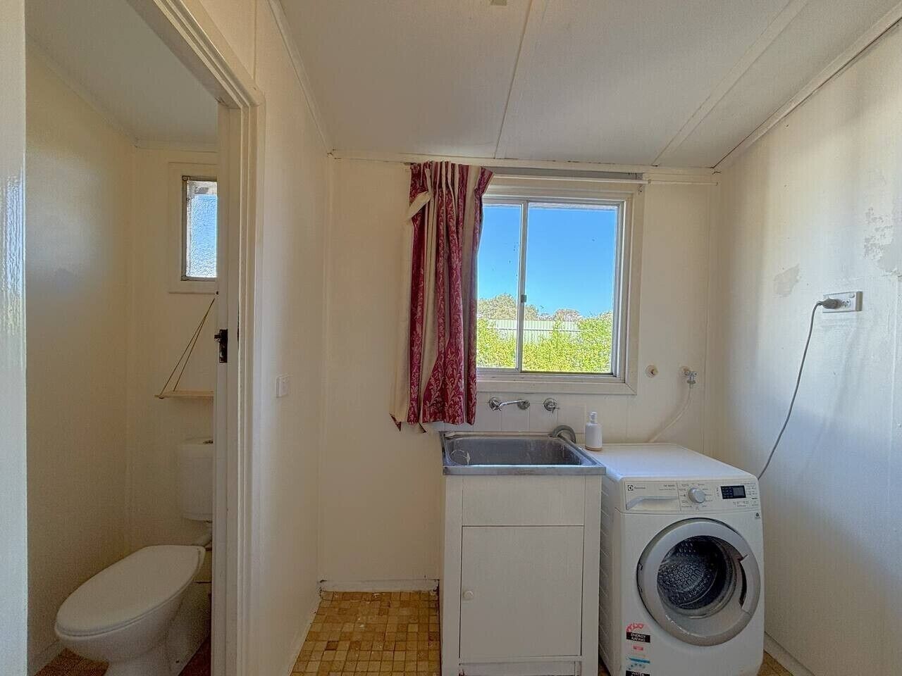 A small laundry room with a washing machine, a sink, and a window with curtains.