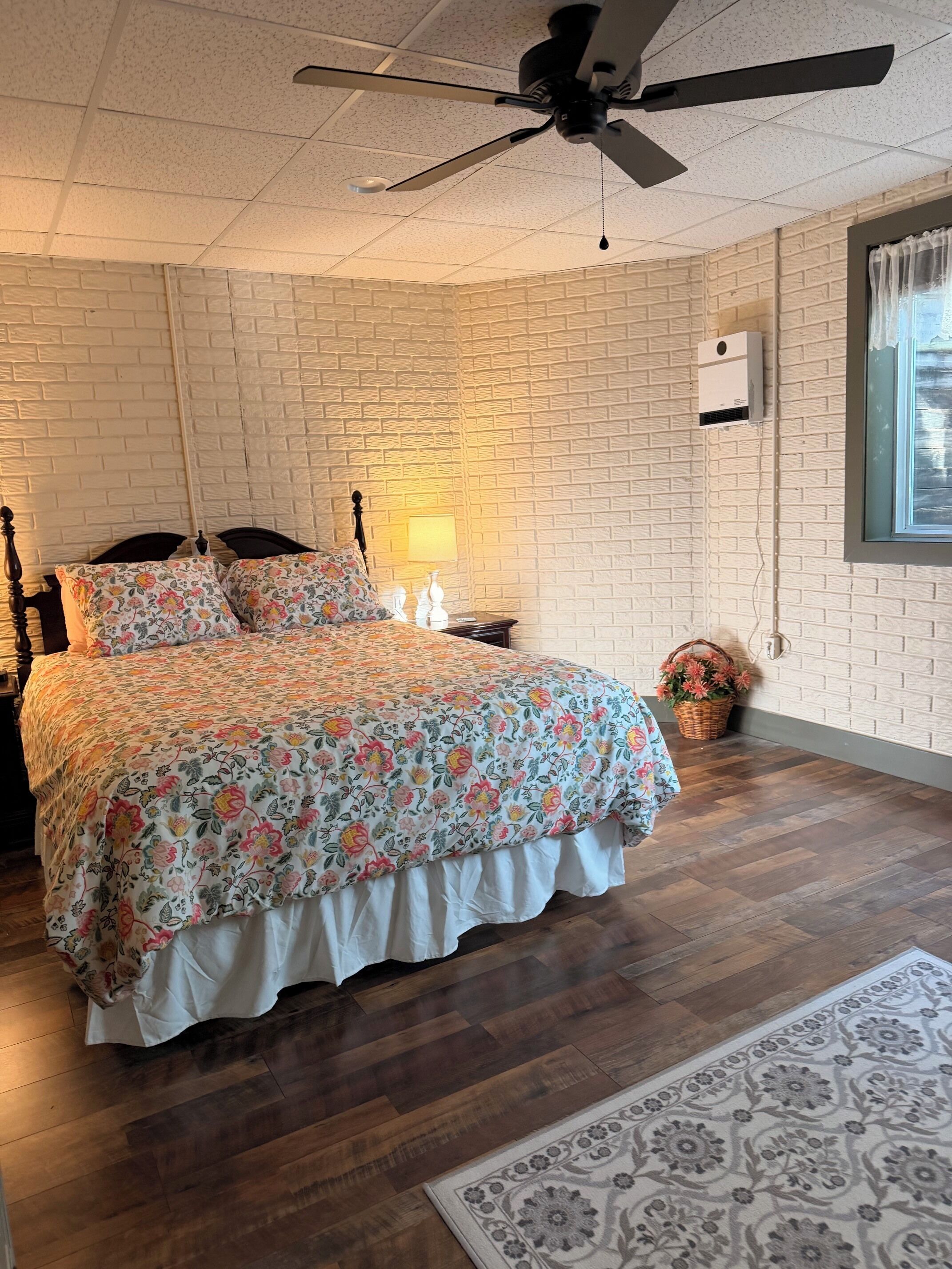 A bedroom with a bed, a ceiling fan, a window, and a brick-patterned wall.
