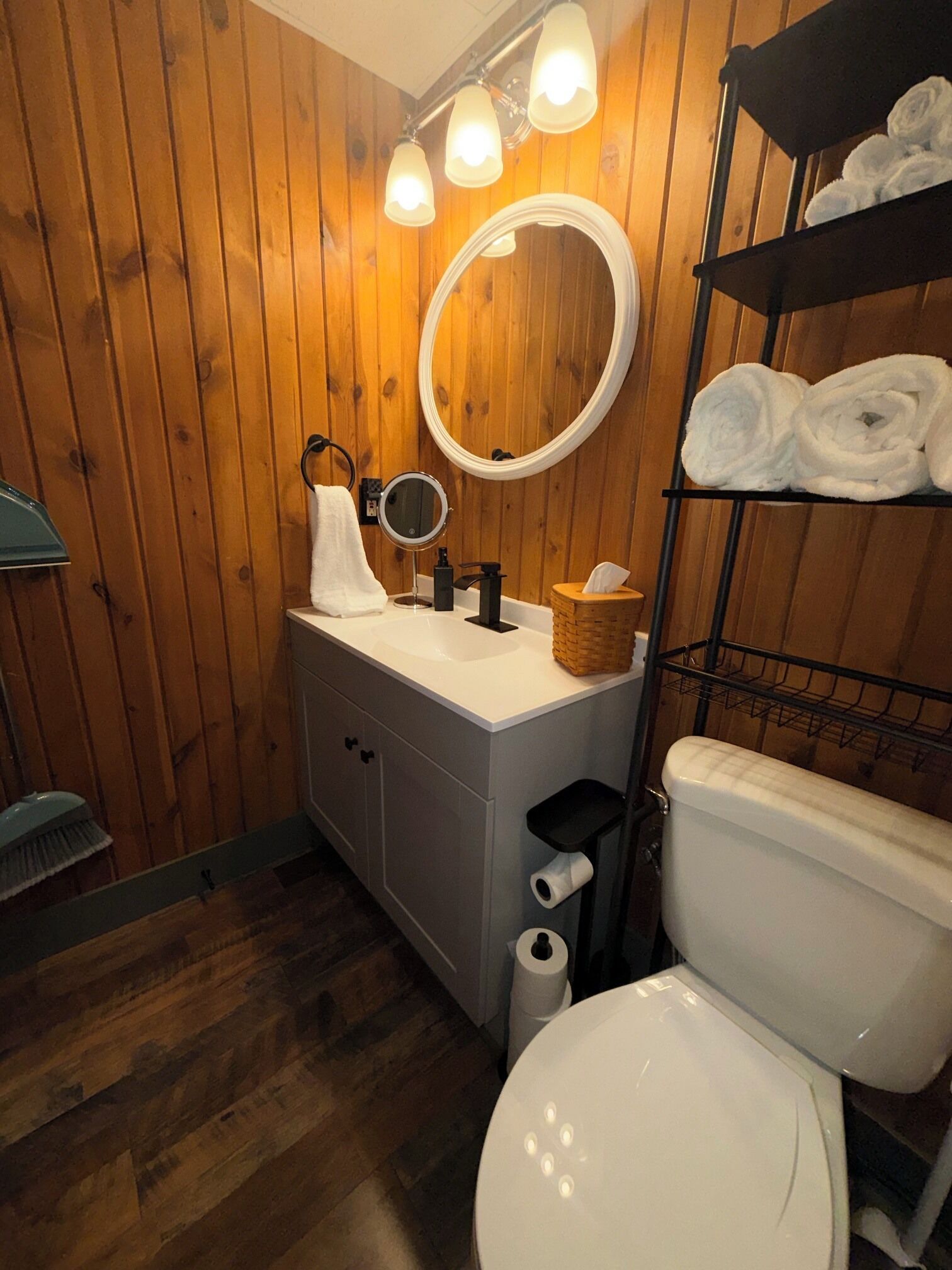 Une salle de bain avec des murs en bois, un lavabo blanc, un miroir rond, des toilettes et une étagère avec des serviettes.
