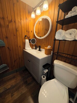Et badeværelse med trævægge, en hvid vask, et rundt spejl, et toilet og en hylde med håndklæder.
