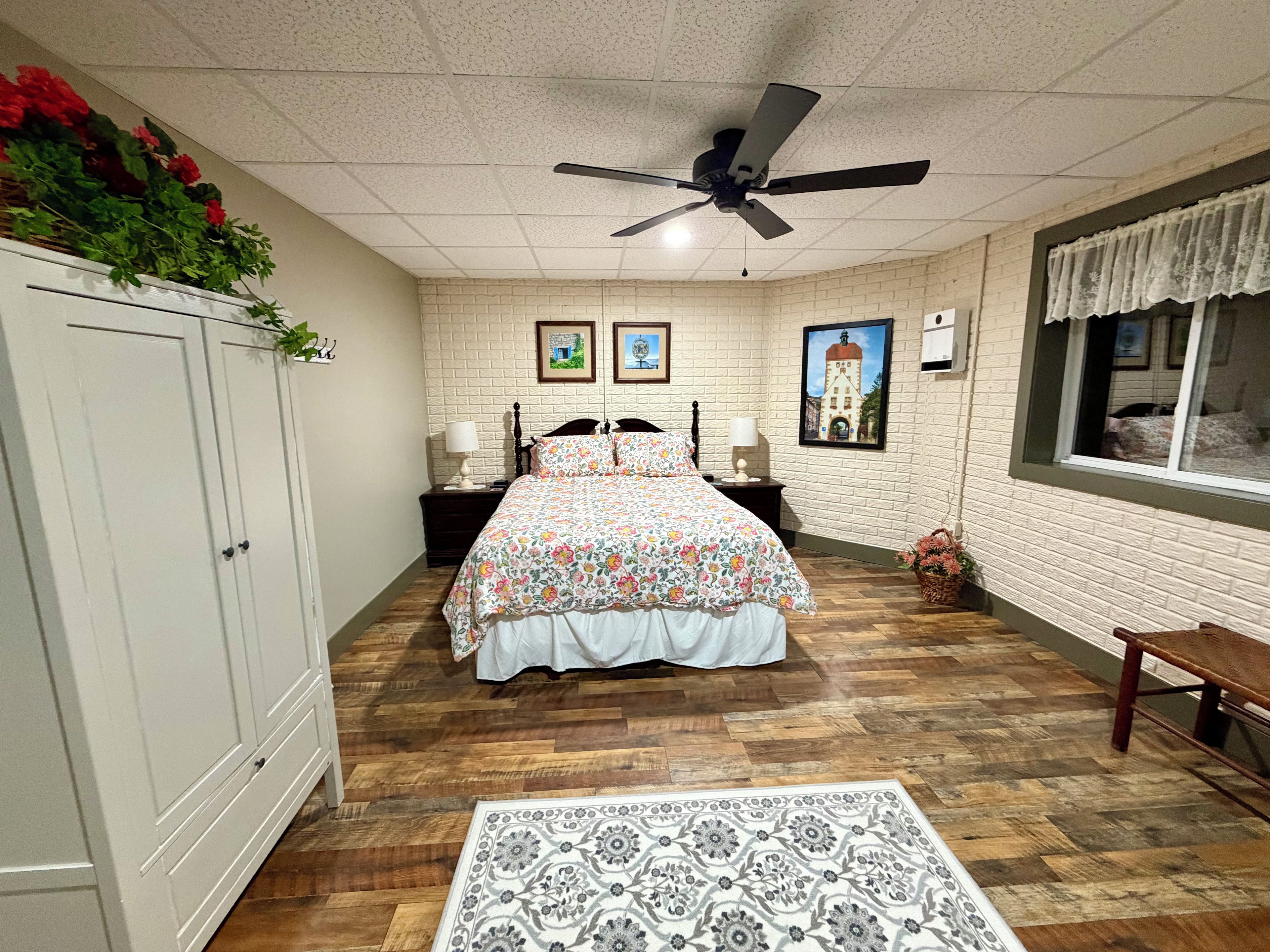 A bedroom with a bed, a ceiling fan, a nightstand, a lamp, a window with a view, and a wall with pictures.