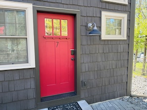 A red door with a number 7, a mailbox, and a small window on a gray building.