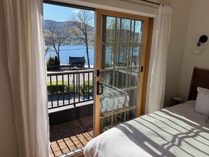 Un dormitorio con cama, mesita de noche, ventana con vistas a un lago y montañas, y balcón con barandilla.