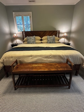 Une chambre avec un grand lit, deux tables de chevet avec des lampes, un banc en bois, une fenĂȘtre avec des stores et un sol Ă motifs.