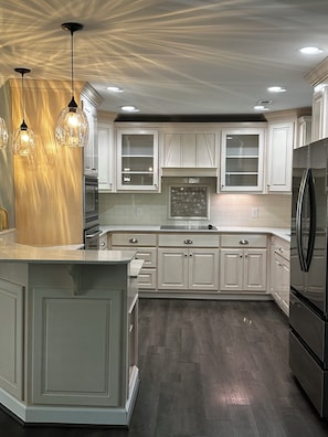 Une cuisine moderne avec des meubles blancs, un ßlot central, des luminaires suspendus et un réfrigérateur en acier inoxydable.