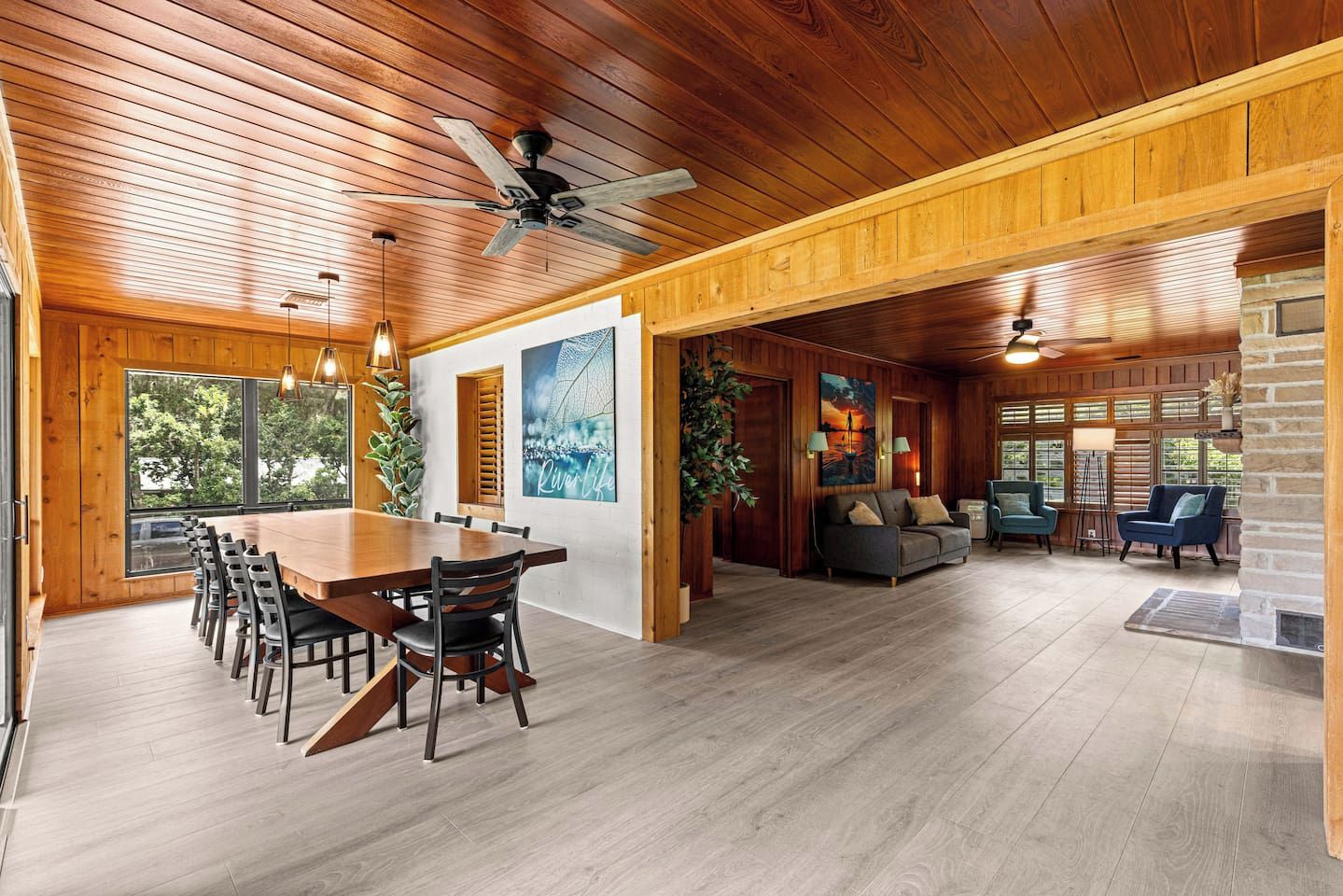 A spacious room with a wooden ceiling, a dining area with a long table and chairs, a seating area with sofas and a fireplace, and large windows allowing natural light.