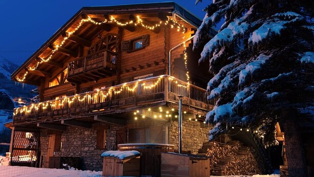 Een houten chalet met een stenen fundering, een balkon met lichtjes en een besneeerd landschap.