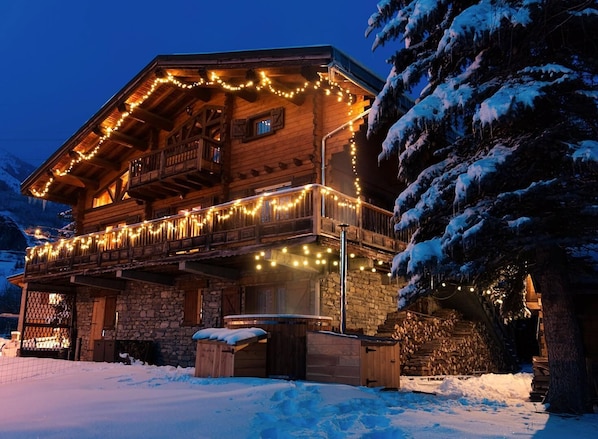A wooden chalet with a stone foundation, a balcony with string lights, and a snow-covered landscape. - The Chalet Des Sangliers (SEEZ)