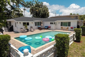 Une piscine avec des bouées gonflables, un patio couvert avec des chaises, et un bâtiment à deux étages avec un toit plat.
