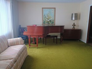 Ein Zimmer mit Klavier, Sofa, Schreibtisch, Lampe und einem Bild an der Wand.