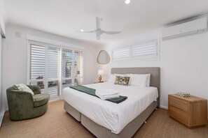 Ein Schlafzimmer mit Bett, Stuhl, Nachttisch, Deckenventilator und Fenster mit Jalousien.