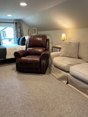 Une chambre d’hôtel avec un fauteuil en cuir marron, un canapé beige, une lampe et un lit donnant sur l’extérieur.