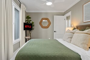 Ein Schlafzimmer mit einer grün gesteppten Bettdecke, einem Wandfernseher, einem sechseckigen Spiegel und einer Tür mit weißem Rahmen.