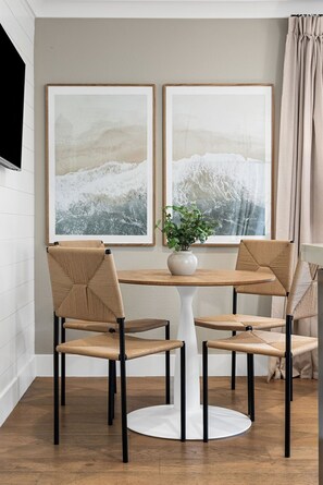 Ein Essbereich mit einem runden Tisch, vier gewobenen Stühlen, zwei gerahmten Bildern an der Wand, einer Zimmerpflanze und einem Fenster mit Vorhängen.