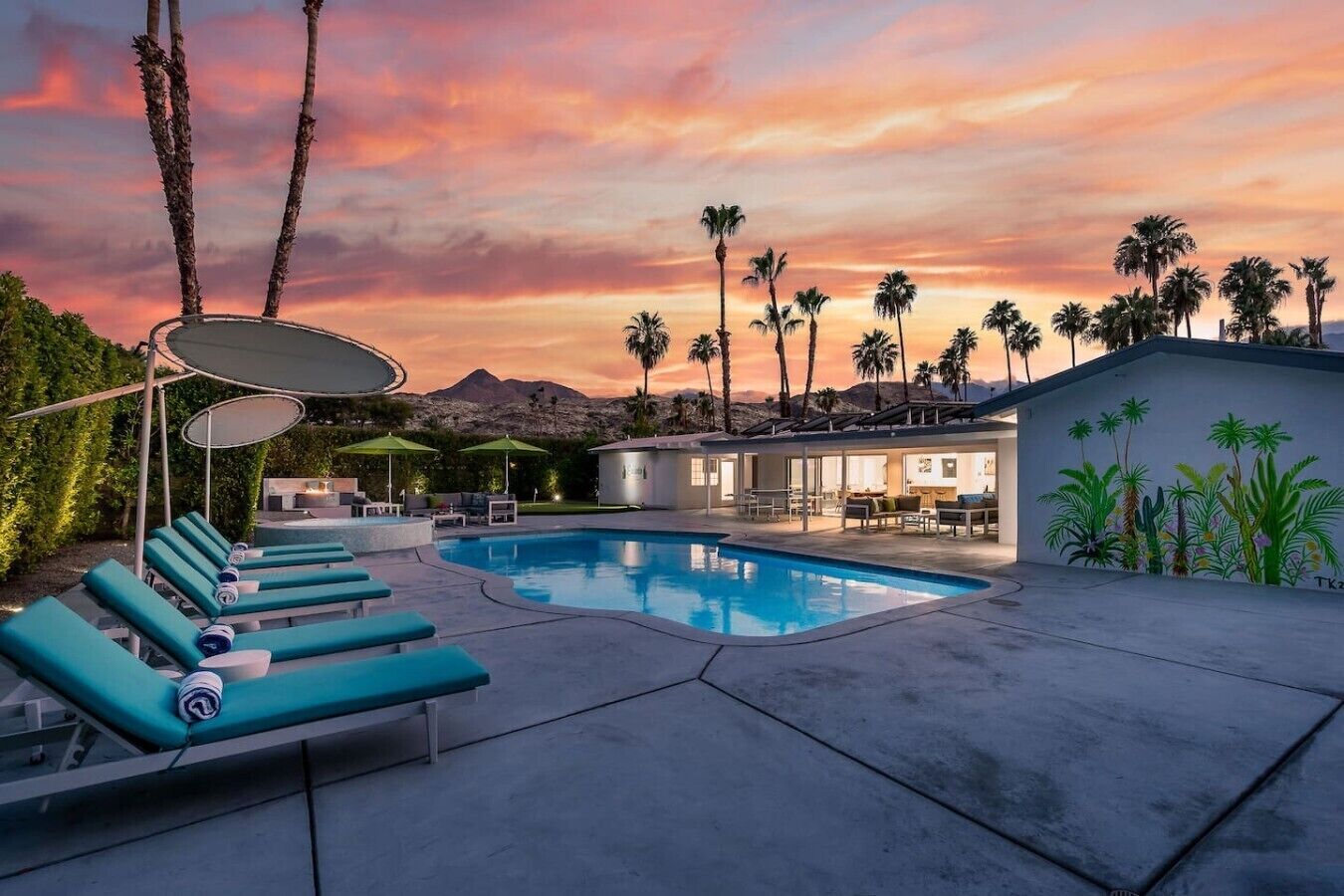 A pool area with lounge chairs, umbrellas, and a view of palm trees and mountains.
