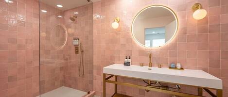 Une salle de bain moderne avec une douche à parois de verre, un meuble-lavabo blanc doté de robinetterie dorée et des murs recouverts de carreaux roses.