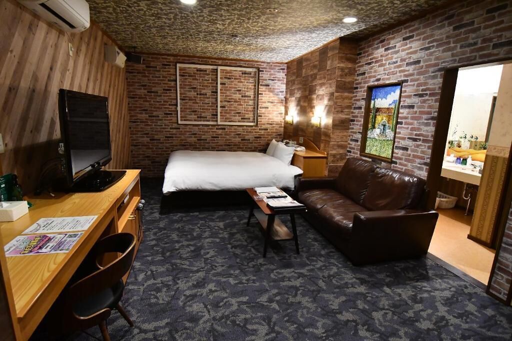 A hotel room with a bed, a desk with a computer, a leather sofa, a small table, and a TV.