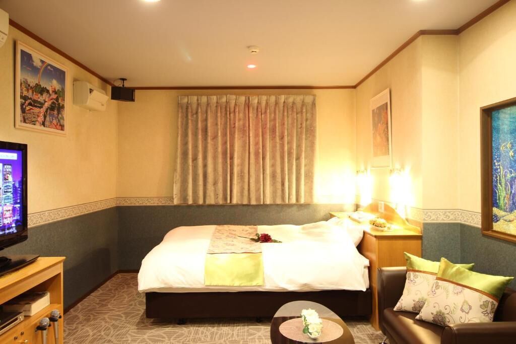 A hotel room with a bed, bedside table, lamp, TV, and a small round table with a flower vase.