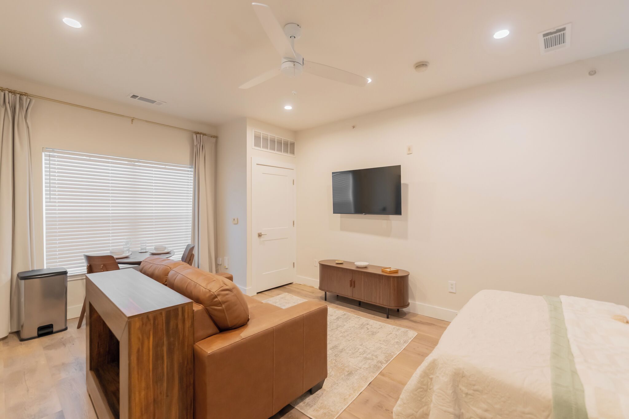 A modern hotel room with a sofa, a TV, a bed, a dining area, and a ceiling fan.