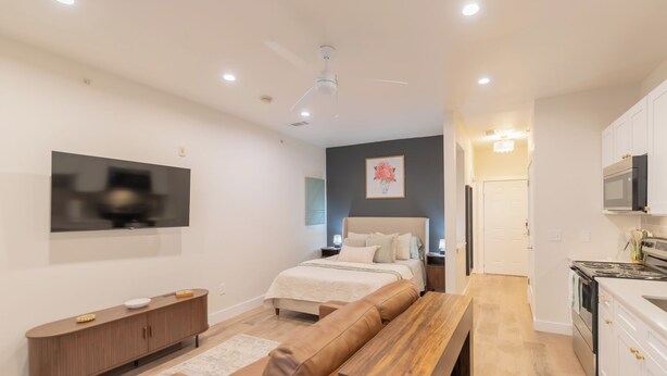 A modern bedroom with a bed, bedside tables, a TV, a wooden bench, and a kitchen area with cabinets and appliances.