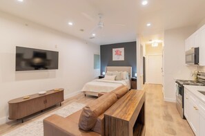 Ein modernes Schlafzimmer mit Bett, Nachttischen, einem Fernseher, einer hölzernen Bank und einer Küchenzeile mit Schränken und Geräten.