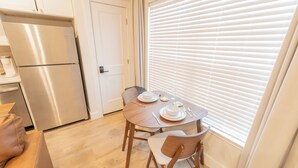 A compact kitchen with a stainless steel refrigerator, a dining table set with white plates and cutlery, and a window with blinds.