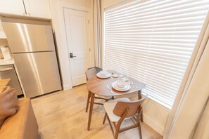 Una cocina compacta con un refrigerador de acero inoxidable, una mesa de comedor con platos y cubiertos blancos, y un ventanal con persianas.